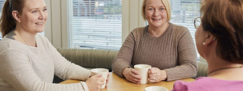 Kolme naista juo kahvia ja keskustelee pöydän ääressä. Naisilla kahdesta on kuulolaitteet ja yhdellä sisäkorvaistute.