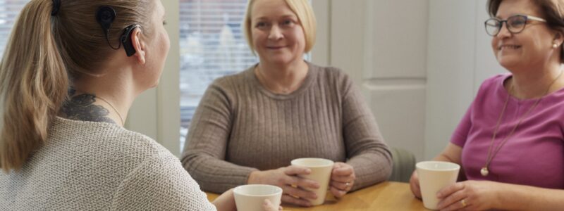 Kolme naista juo kahvia ja keskustelee pöydän ääressä. Kahdella naisista on kuulokoje ja yhdellä sisäkorvaistute.